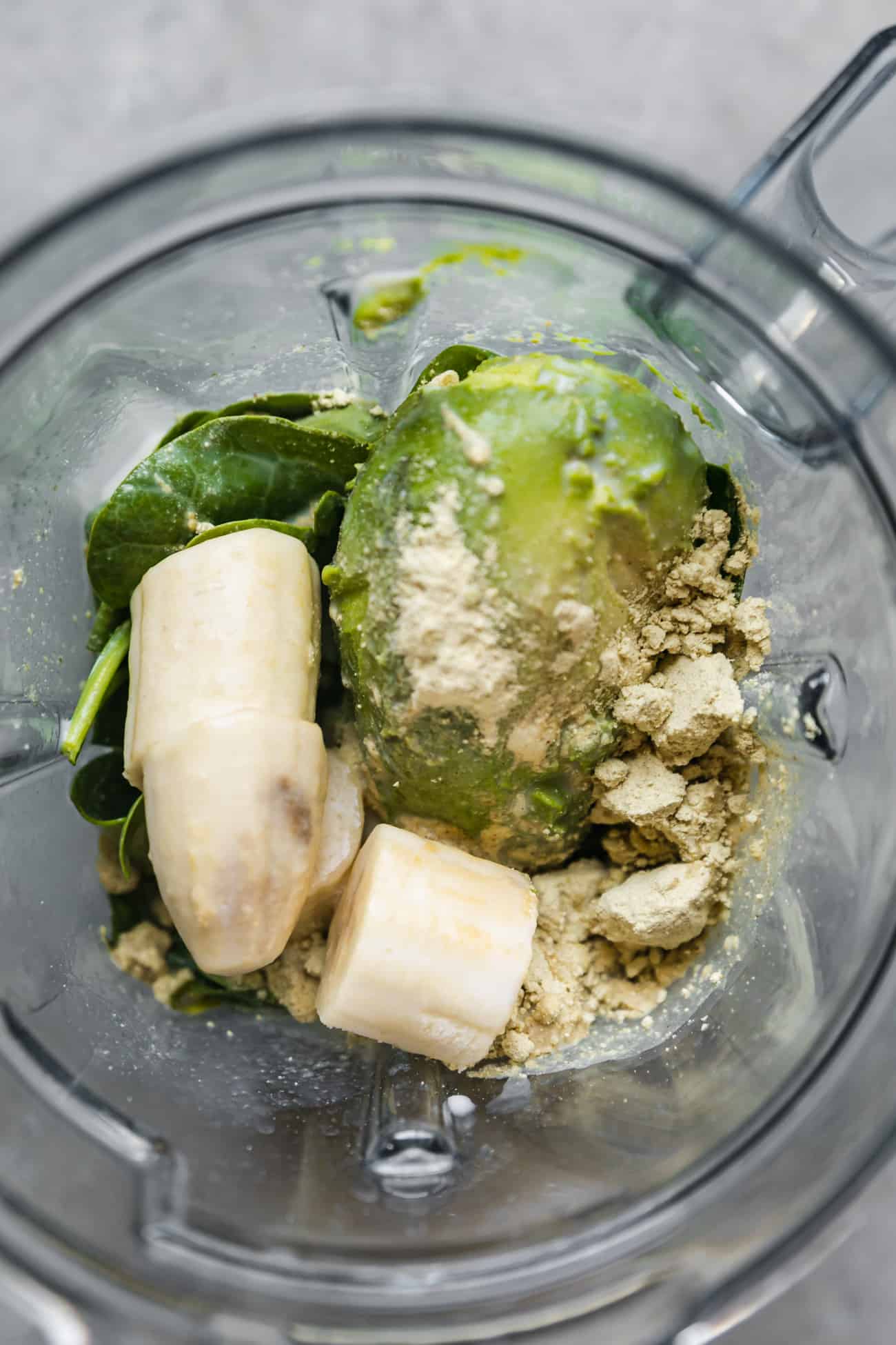 ingredients dumped into a blender for a creamy avocado smoothie: frozen banana, avocado, spinach, protein powder.